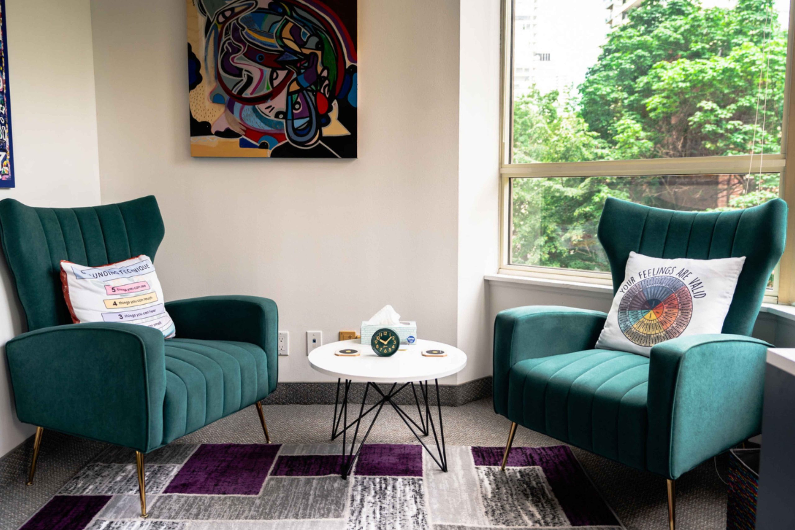 two green lounge chairs on each side of the frame with decorative pillows pm each one pillow says your feelings are valid. there is a small round table in betweent he two chairs with a box of tissue and a green clock.